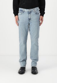 Világoskék denim farmerek egyenes szabással, öt zsebbel és gombos záródással. Fekete cipővel és fekete hosszú ujjú inggel viselve.