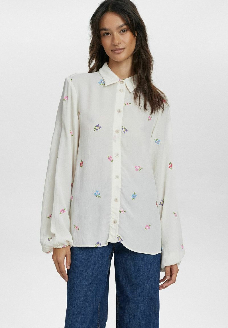 Camicia bianca con ricami floreali in rosa, blu e verde. Presenta un colletto classico e maniche lunghe e voluminose. Abbinata a jeans blu.