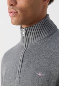 Nahaufnahme einer Person, die einen grauen, gerippten Rollkragenpullover mit einem kleinen gestickten roten, weißen und blauen Schild-Logo auf der Brust trägt.