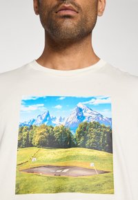 Cream cotton t-shirt featuring a vibrant landscape print of mountains, trees, and a basketball court, showcasing blue skies and green hills.