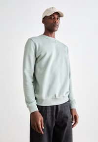 Man in een lichtgroene sweatshirt, donkere jeans en een beige baseballcap, staande tegen een eenvoudige witte achtergrond.