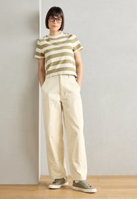 T-shirt de mangas curtas listrado em tons de oliva e creme com um corte descontraído, combinado com calças largas creme e sapatos verdes. Fundo de superfície lisa.