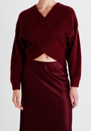 Femme portant un pull cache-cœur duveteux rouge foncé découvrant le ventre et une jupe en satin brillant assortie.