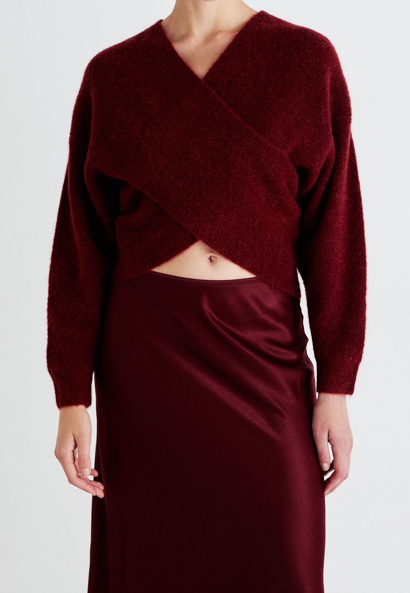 Femme portant un pull cache-cœur duveteux rouge foncé découvrant le ventre et une jupe en satin brillant assortie.