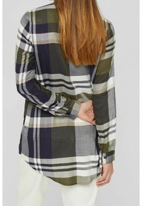 Personne aux longs cheveux bruns portant une grande chemise à carreaux verte, noire, grise et blanche, une main derrière le dos.