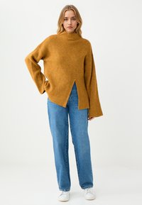 Musztardowy żółty sweter typu turtleneck z prążkowanego materiału, z wysokim dekoltem, detalem w postaci rozcięć po bokach oraz szerokimi rękawami, w połączeniu z jasnoniebieskimi spodniami prostymi.
