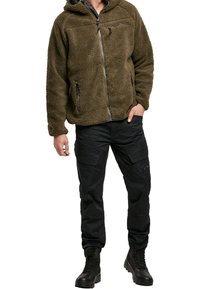 Giacca in fleece verde oliva, con zip davanti e tasche, abbinata a pantaloni cargo neri con più tasche e tessuto strutturato, indossata con stivali neri.
