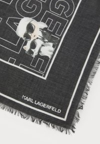Musta huivi, jossa on sarjakuvakuva auringonlaseja käyttävästä hahmosta ja koirasta, valkoisilla rajoilla reunustettuna. Sisältää "KARL LAGERFELD" -merkin.