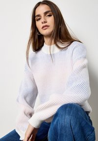 Ribbelgebreide trui in het wit met pasteltinten roze en blauw. Beschikt over een hoge halslijn en lange, volumineuze mouwen. Gecombineerd met blauwe jeans.