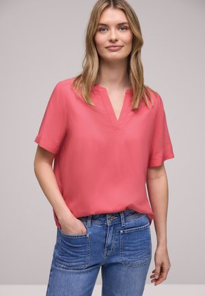 Jeune femme aux cheveux blonds portant un chemisier corail à manches courtes et un jean bleu, debout avec une main dans la poche devant un fond neutre.