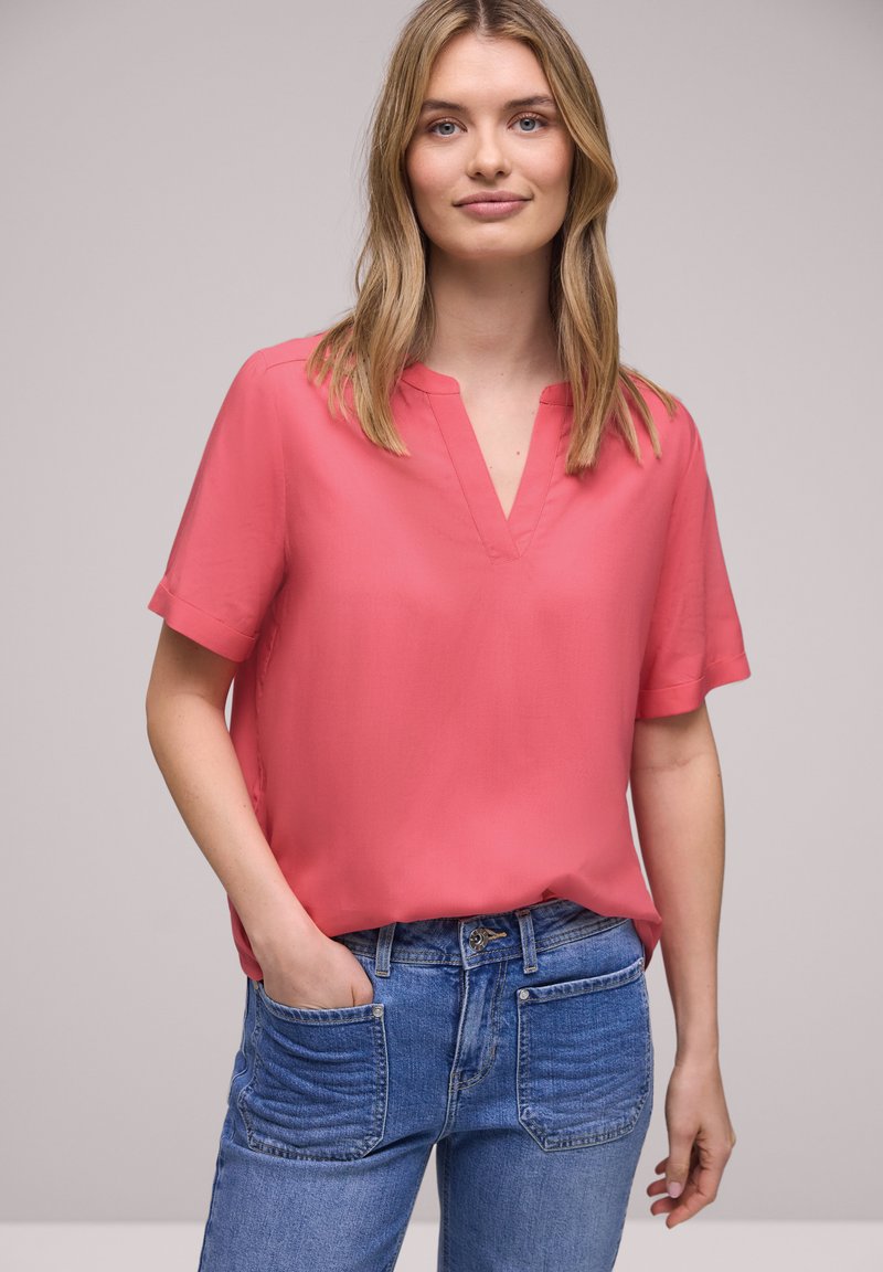 Jeune femme aux cheveux blonds portant un chemisier corail à manches courtes et un jean bleu, debout avec une main dans la poche devant un fond neutre.