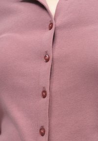 Κοντινό πλάνο dusty rose ζακέτας με κουμπιά. Τα μικρά, στρογγυλά κουμπιά είναι στο ίδιο χρώμα και το ύφασμα είναι πλεκτό με ρίγες.