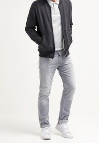 Homme portant une veste noire zippée sur un polo gris, jean gris clair avec les revers roulés, et des baskets blanches, debout les mains dans les poches.