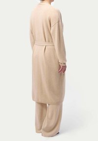 Beige stickad lång kappa med vida ärmar och en knytbälte, matchad med löst sittande byxor. Mjuk textur, enkel design.