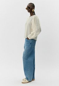 Beige lätt genomskinlig blus med ögla mönster, puffärmar, kombinerad med lösa blå denimjeans. Modellen bär runda solglasögon och beiga skor.