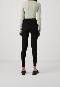 Woman wearing black high-waisted skinny jeans and a light green ribbed long-sleeve top, standing on a plain white floor and wall.