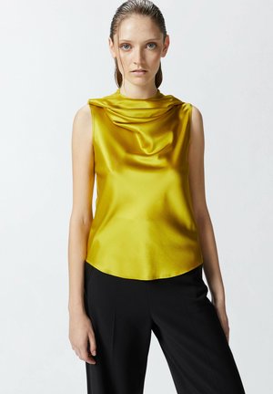 Top sin mangas de satén amarillo con escote drapeado y silueta fluida, combinado con pantalones negros, exhibido contra un fondo claro.