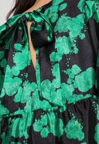 Abito in tessuto di seta nera con ampia stampa floreale verde smeraldo, caratterizzato da un fiocco annodato al collo e un'apertura a chiave sotto il fiocco.