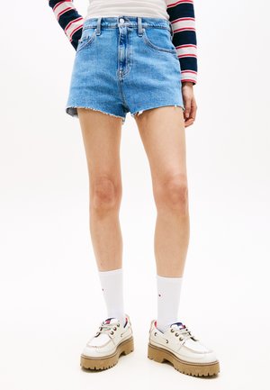 Persona che indossa pantaloncini di jeans a vita alta blu, calzini bianchi, scarpe bianche robuste e una camicia a maniche lunghe a righe, con le mani sui fianchi.