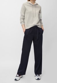 Světle šedá mikina s kapucí a přední kapsou, s třemi bílými pruhy na jednom rukávu, v kombinaci s námořnickými kalhotami širokého střihu a vícebarevnými teniskami.