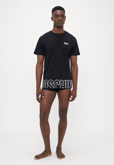T-shirt en coton noir avec un logo "Moschino" blanc sur la poitrine et une impression de logo plus grande à l'ourlet, porté avec un sous-vêtement noir assorti.