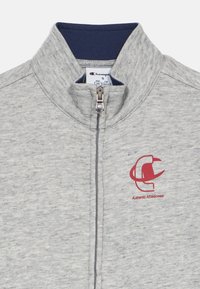 Sweat-shirt à fermeture éclair gris avec un col montant, orné d'une bordure marine et d'un logo rouge. Le tissu présente une texture chinée et une coupe standard.