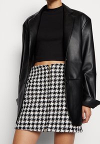 Blazer surdimensionné en cuir noir porté sur un haut noir ajusté, associé à une mini-jupe taille haute à motif pied-de-poule avec une fermeture éclair à l'avant.