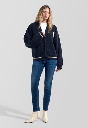 Mujer con un cárdigan oscuro de gran tamaño, vaqueros ajustados azules, botines blancos y una blusa blanca, de pie sobre un fondo gris liso.