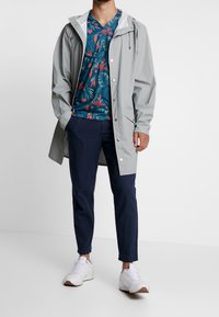 Impermeable gris con botones a presión, camisa floral en tonos azul verdoso, pantalones navy y zapatillas blancas con suelas marrones. Un atuendo simple y funcional para un uso casual.