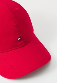 Casquette de baseball rouge en tissu texturé, avec une visière incurvée et un petit logo comportant des rayures noires, blanches et rouges sur le côté.