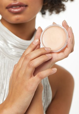 Genomskinlig plastbehållare som innehåller ett ljusbeige balsam, hålls i en hand med naturliga naglar. Bakgrunden är neutral med fokus på produkten.