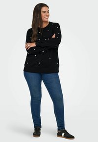 Schwarzer Sweatshirt mit kleinen rosa Herzmustern, runder Halslinie und langen Ärmeln, kombiniert mit blauen Skinny-Jeans und schwarzen Sneakers.