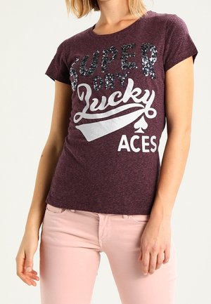 Femme portant un T-shirt bordeaux avec le texte et les graphiques "Super Dry Lucky Aces", associé à un pantalon rose clair sur un fond uni.