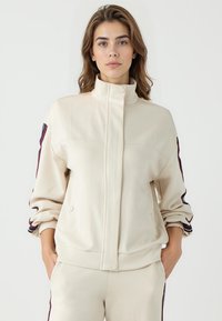 Beige Zip-Jacke mit hohem Kragen, Seitentaschen und aufgerollten Ärmeln. Verfügt über kontrastierende Streifen entlang der Schultern in Dunkelrot und Blau.