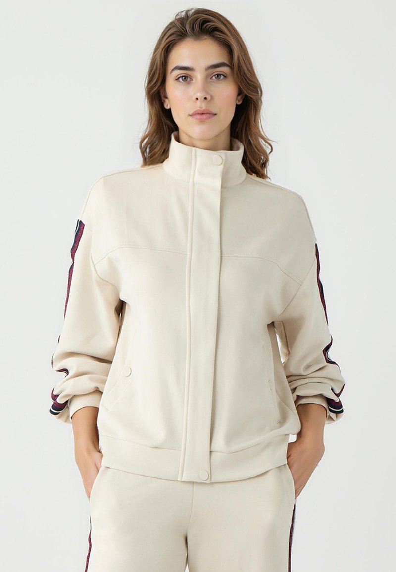 Beige Zip-Jacke mit hohem Kragen, Seitentaschen und aufgerollten Ärmeln. Verfügt über kontrastierende Streifen entlang der Schultern in Dunkelrot und Blau.
