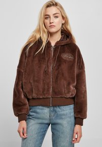 Hoodie en fausse fourrure marron avec fermeture éclair à l'avant, poignets et ourlet côtelés, et un détail de logo brodé sur la poitrine. Porté avec un jean bleu clair.