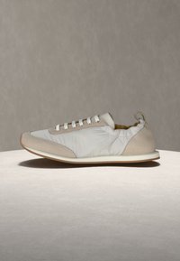 Μπεζ και λευκό αθλητικό παπούτσι με λευκά κορδόνια και χαμηλή σόλα σε ουδέτερη επιφάνεια και φόντο.