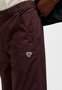Bordeauxrode broek met een elastische tailleband, voorzien van zijzakken en een geweven logopatch in crèmekleur op het bovenbeen. Gladde stoftextuur.