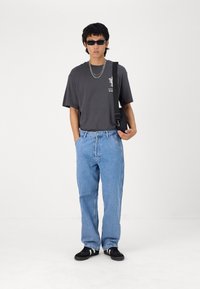 Grå kortermet t-skjorte med grafisk trykk, løse blå jeans, svarte joggesko og mørke solbriller. Modellen har på seg en sølvkjede og veske.