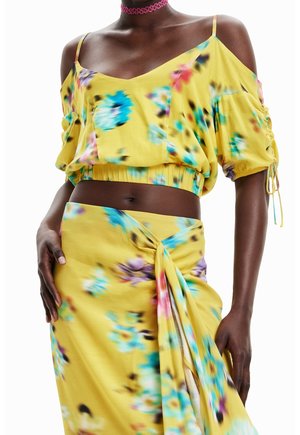 Mørkhudet kvinde iført en gul blomsterprintet cold-shoulder crop top og matchende omslagsskjørt, accessoriseret med en pink choker-halskæde.