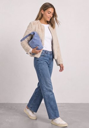 Kvinde går iført beige pelsjakke, hvid skjorte, blå jeans og hvide sneakers, mens hun holder en stor blå håndtaske på armen.