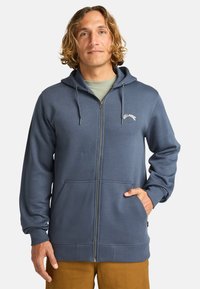 Blå zip-up hoodie lavet af blødt stof, med en kængurulomme, snoretræk i hætten og et logo på brystet. Ribstrikkede manchetter og kant.