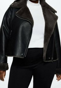 Vrouw in een zwart faux leren jack met donkere bontvoering, een witte geribbelde top en zwarte broek tegen een effen achtergrond.