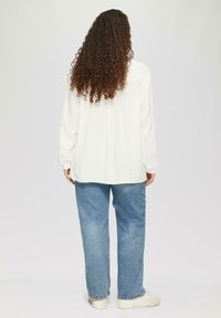 Witte, gestructureerde oversized shirt met lange mouwen, gecombineerd met lichtblauwe relaxed-fit jeans en witte sneakers, gezien vanuit de rug.