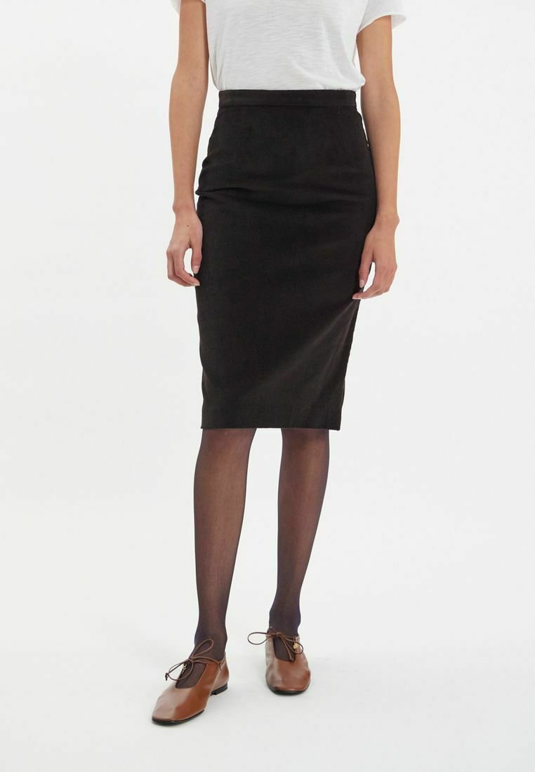 Jupe crayon noire en tissu doux, taille haute, longueur genou, associée à une chemise blanche et des chaussures marron, avec des collants sombres.