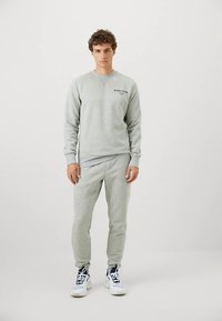 Jovem em pé, vestindo um sweatshirt cinza claro e calças de jogging, com tênis brancos e azul-marinho, contra um fundo simples.