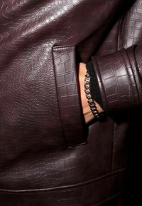 Donker bordeaux leren jack met een gestructureerd, reliëfpatroon. Voorzien van een zijzak en een aansluitende manchette. Zilveren kettingarmband zichtbaar op de hand.