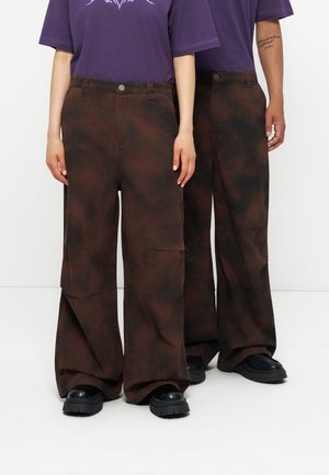 Deux personnes portant un pantalon marron à jambes larges, des chaussures noires et des t-shirts violets, debout côte à côte sur un fond blanc.