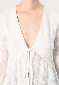 Weißes Bluse mit tiefem V-Ausschnitt, floral strukturiertem Stoff, langen Ärmeln und einem Bindedetail am Ausschnitt. Helles und zartes Material.