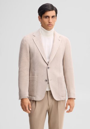 Uomo con blazer beige chiaro a spina di pesce sopra dolcevita bianco e pantaloni beige, in piedi davanti a uno sfondo chiaro e uniforme.